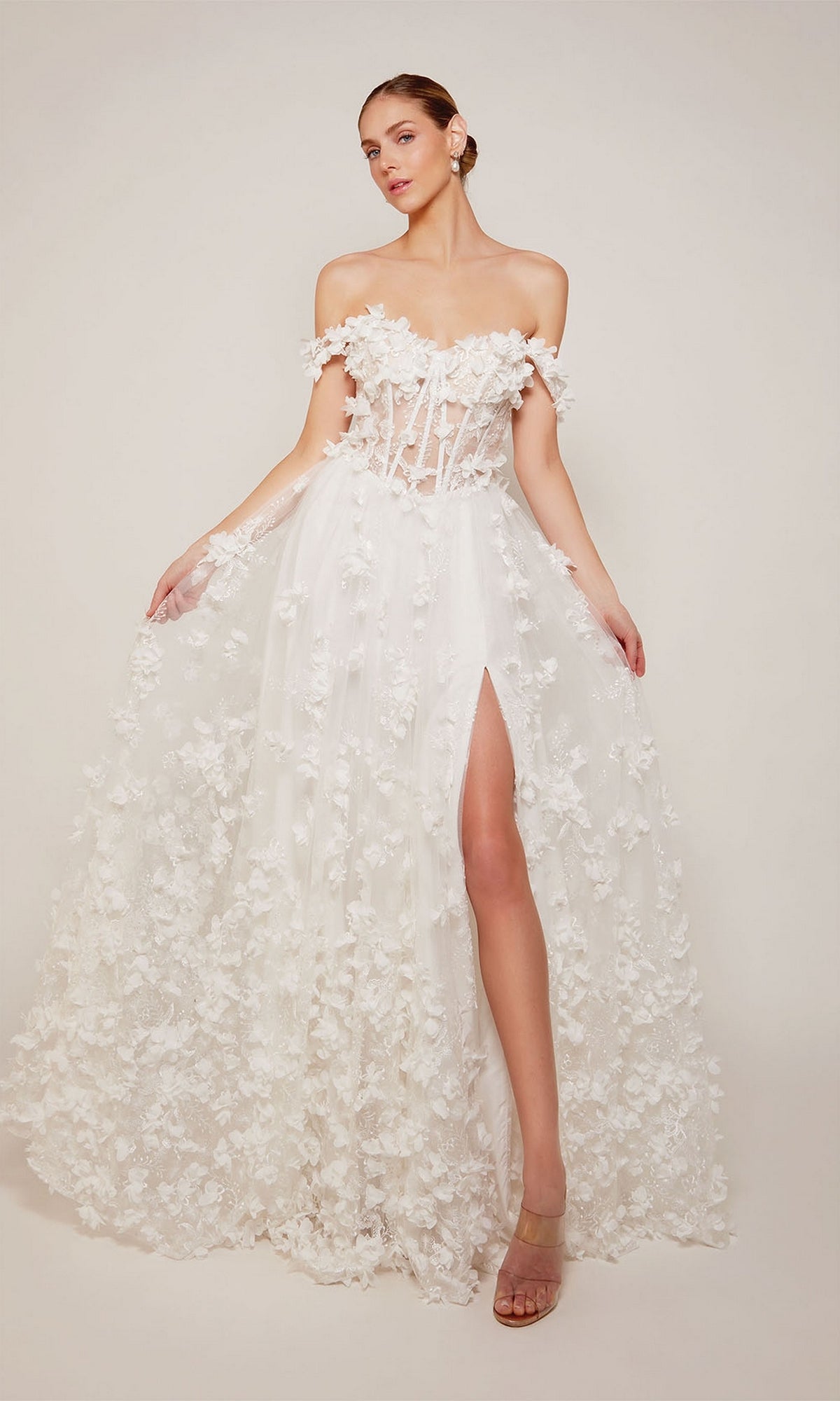 A woman models the Alyce 61308 Off-Shoulder Long Floral Dress—an elegant white gown with floral appliqués, sheer bodice, and high slit—posed against a plain backdrop.